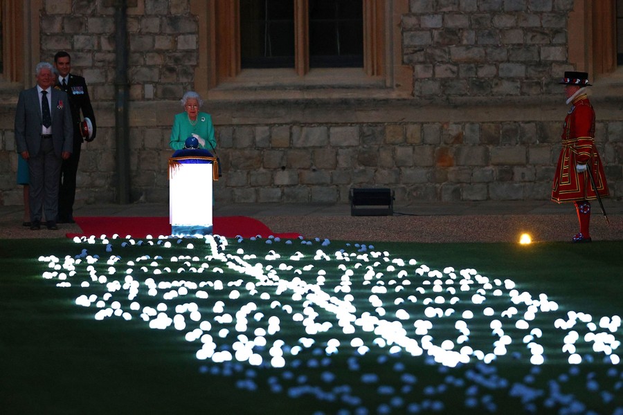 The queen of England stands at an illuminated podium with a long pathway of illuminated lights on the ground in front of her.