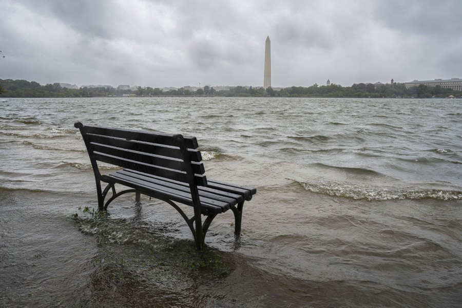 A park bench sits is shallow floodwater on a stormy day, with a view of the Washington Monument.