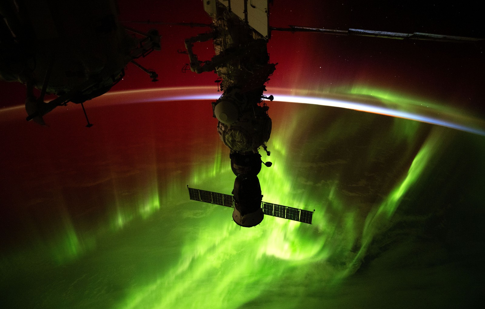 Green and red streaks of light from an aurora appear above the Earth, seen from orbit, with part of the International Space Station visible at center.