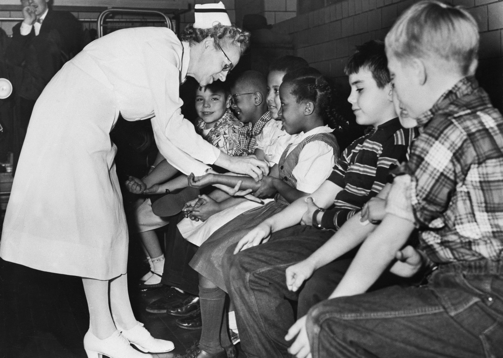 A nurse leans over to give a shot to one of a half-dozen young children seated on a bench.