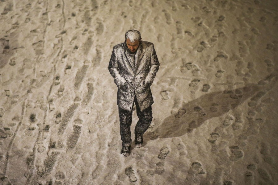 A snow-covered man walks on a snow-covered sidewalk.