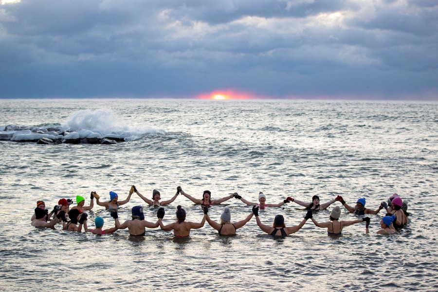 A group of swimmers wearing winter hats hold hands in a circle, chest-deep in a lake.