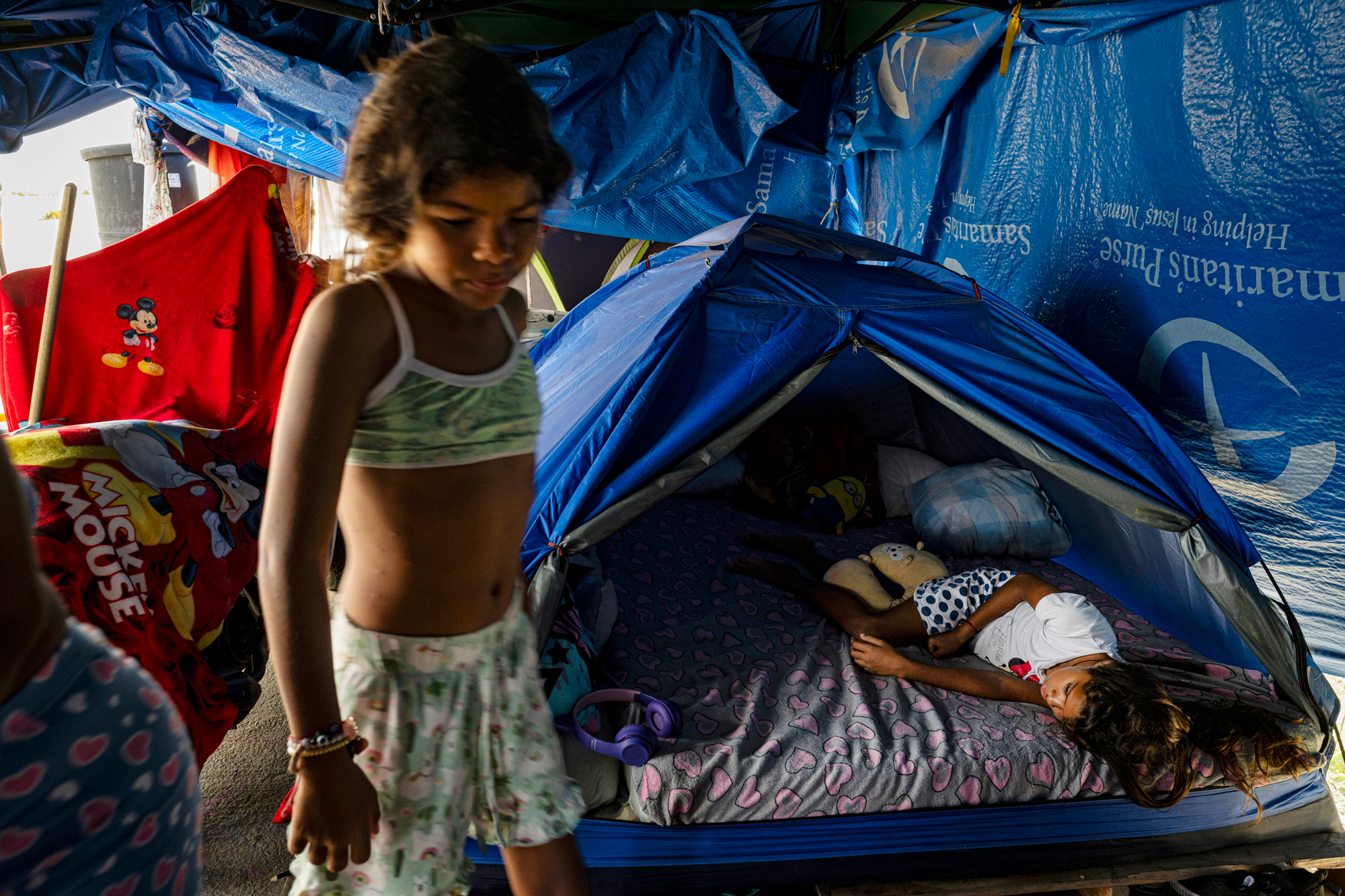 a child sleeps in a blue tent in the background while a child in a green top and shorts stands in the foreground