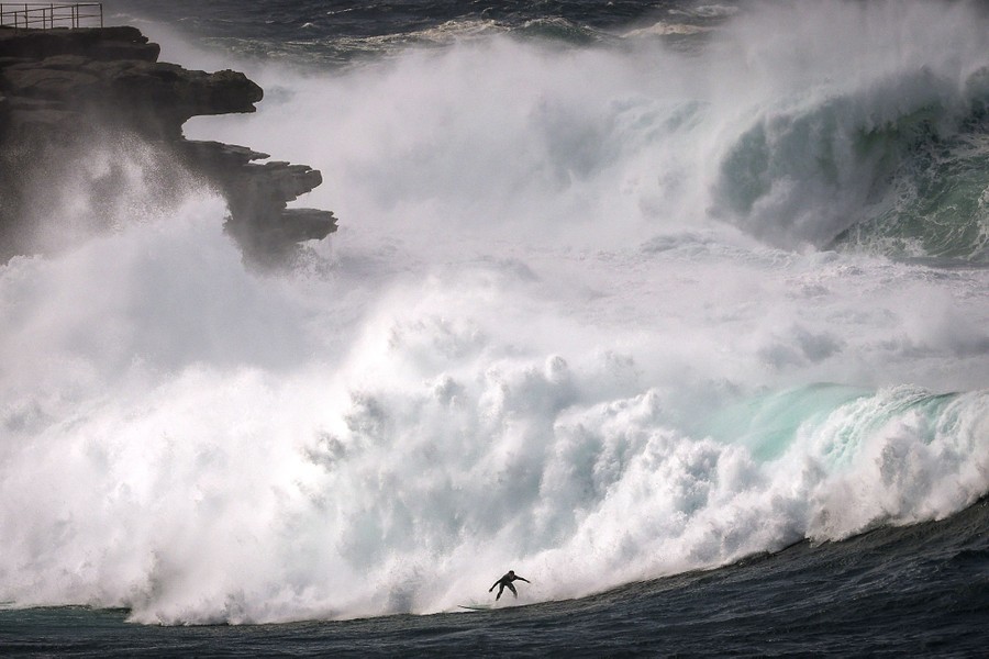 A surfer rides a large wave near a rocky shoreline.