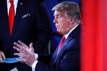 A photo of former President Donald Trump wearing a navy suit and red tie; another man wearing a red tie is in the background holding a stack of papers.