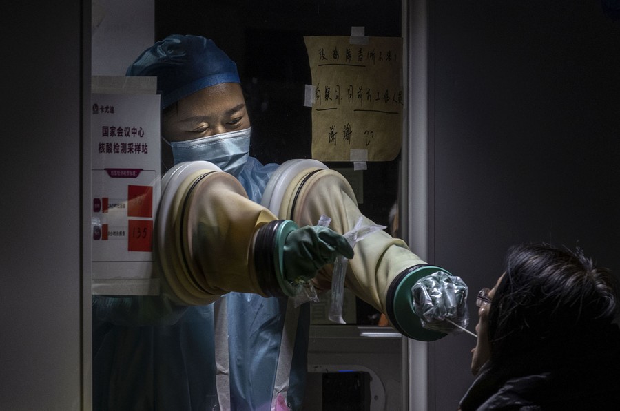 A health-care worker reaches their arms through holes, into rubber gloves, to administer a swab test to a person.