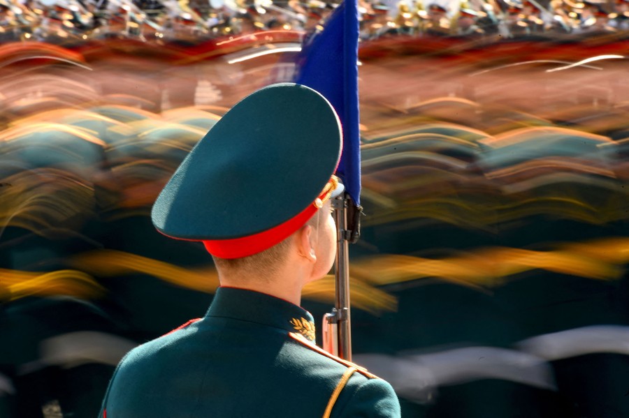 A soldier stands as others march past, seen blurry in a long-exposure image.