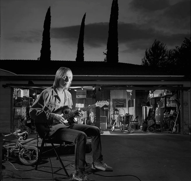 black-and-white photo of Tom Petty sitting in front of garage with open door while playing guitar