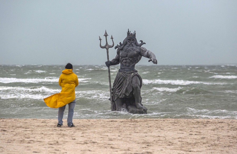 A man looks at a statue of the Greek god Poseidon on a beach on a stormy day.
