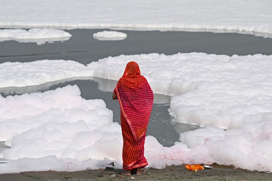 A person stands on the bank of a river that is mostly covered in floating foam.