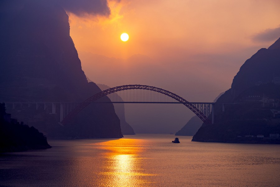A ship sails beneath a road bridge, past steep mountains, at sunrise.