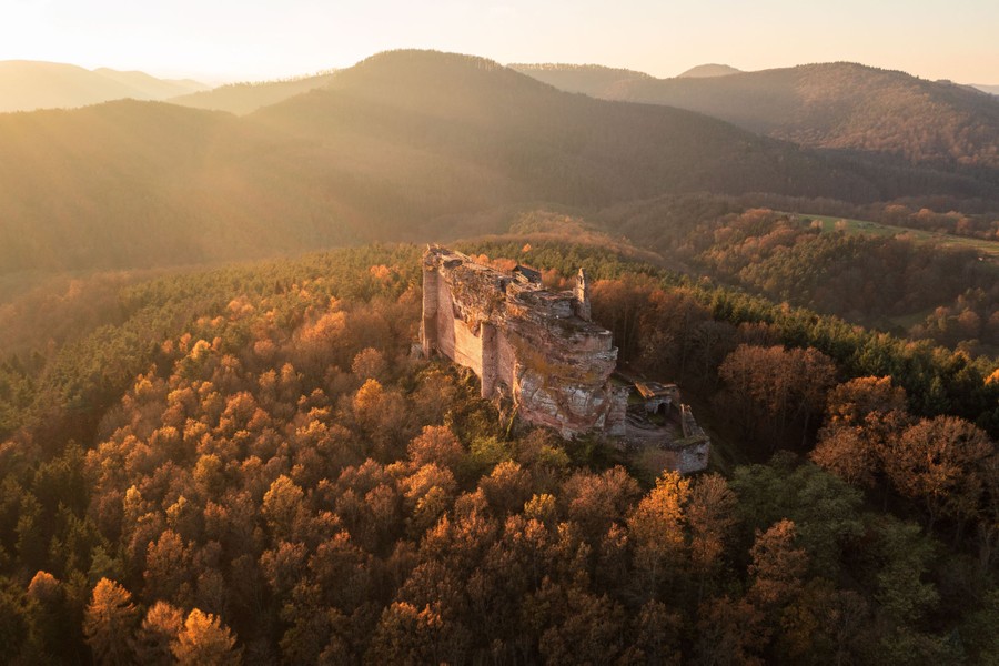 An aerial view of a sunset over forested hills and the ruins of a medieval castle built on a tall rock outcrop.