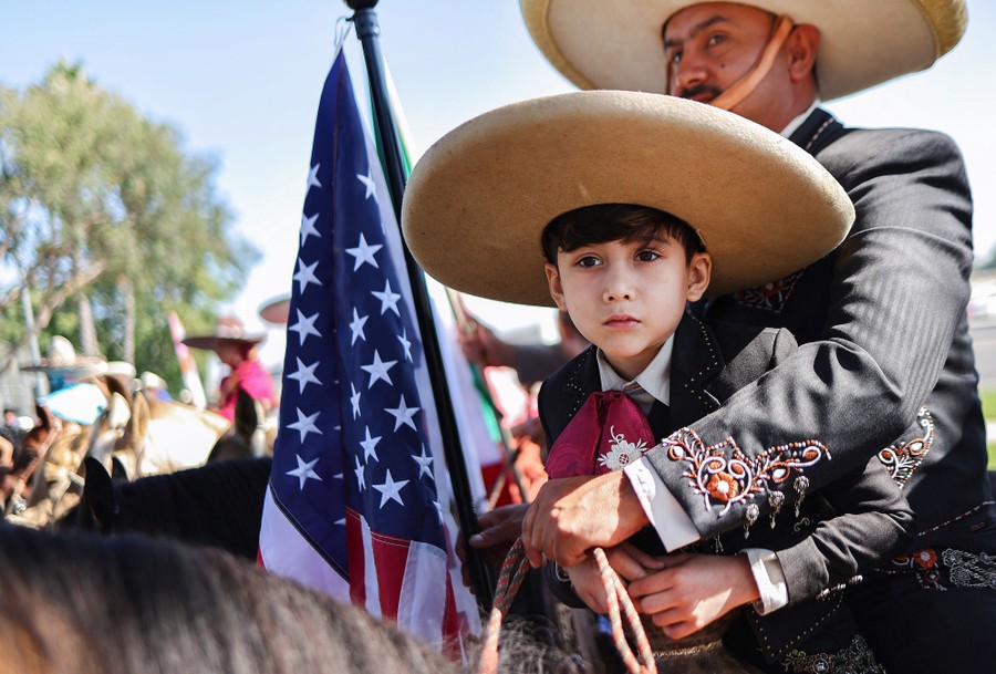 A man and boy sit on horseback together, each wearing embroidered suits and sombreros, while holding an American flag.