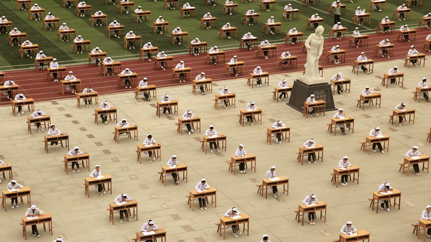 A field filled with desks with test takers at each one, dressed in white and black. The field is green, red, and white.