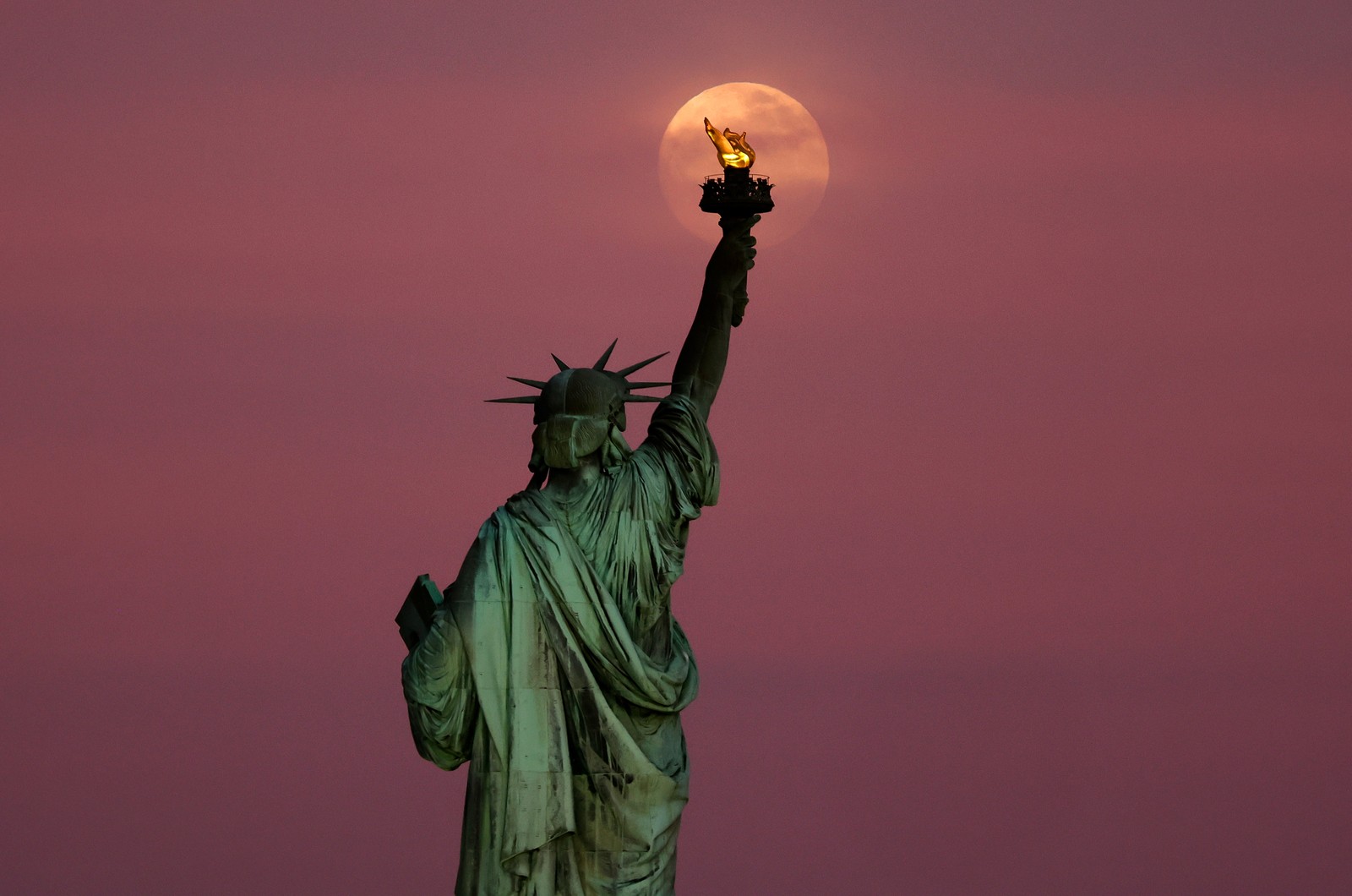 The moon rises, seen through clouds behind the Statue of Liberty.