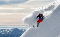 Photograph of a skier in red pants, a blue jacket, and a black helmet carving a turn in deep powder on a steep slope, with mountains in the background