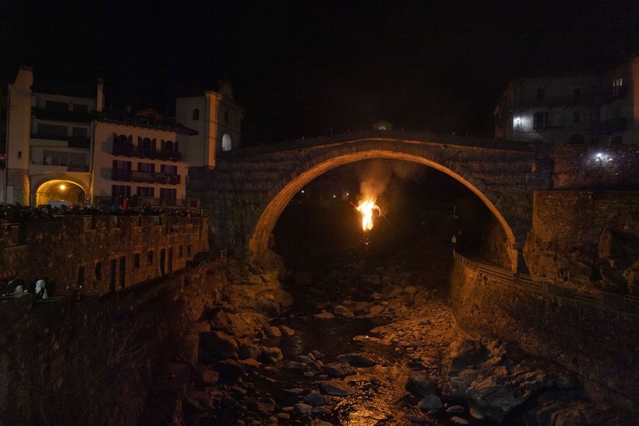 A burning effigy of the devil hangs beneath an old stone bridge over a rocky riverbed in a town.