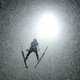 A ski jumper flies through a flurry of snowflakes at night, backlit by a bright light.