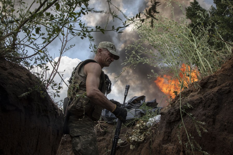 A soldier shelters in a trench as fire and smoke rise nearby.
