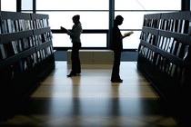 Two women stand next to rows of books, reading. 