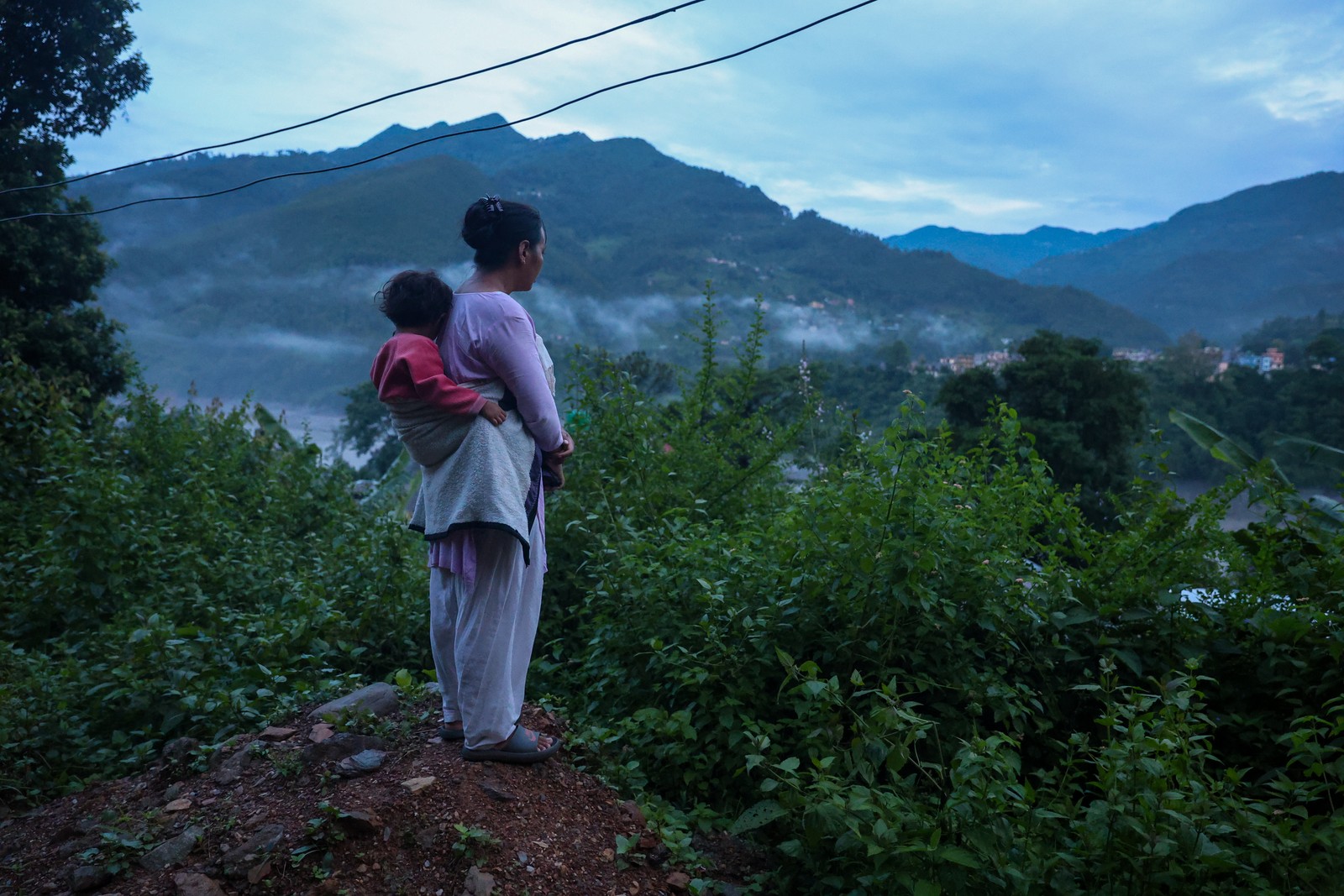 A woman, carrying a small child on her back, looks out over a river valley.