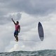 A surfer points upward while jumping above a wave with his surfboard beside him, seeming to stand casually upright in mid-air .