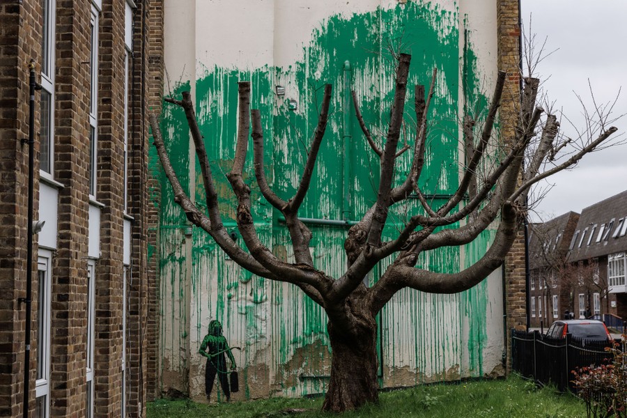 A mural of splashed green paint sits on a wall behind a tree with bare branches.