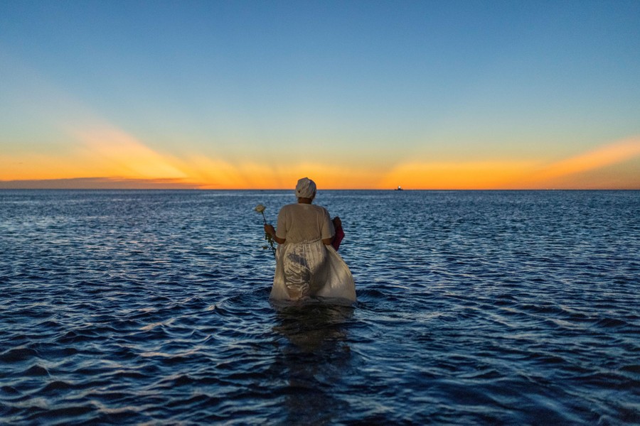 A woman wades into knee-deep water for a religious ritual against a stunning sunrise or sunset