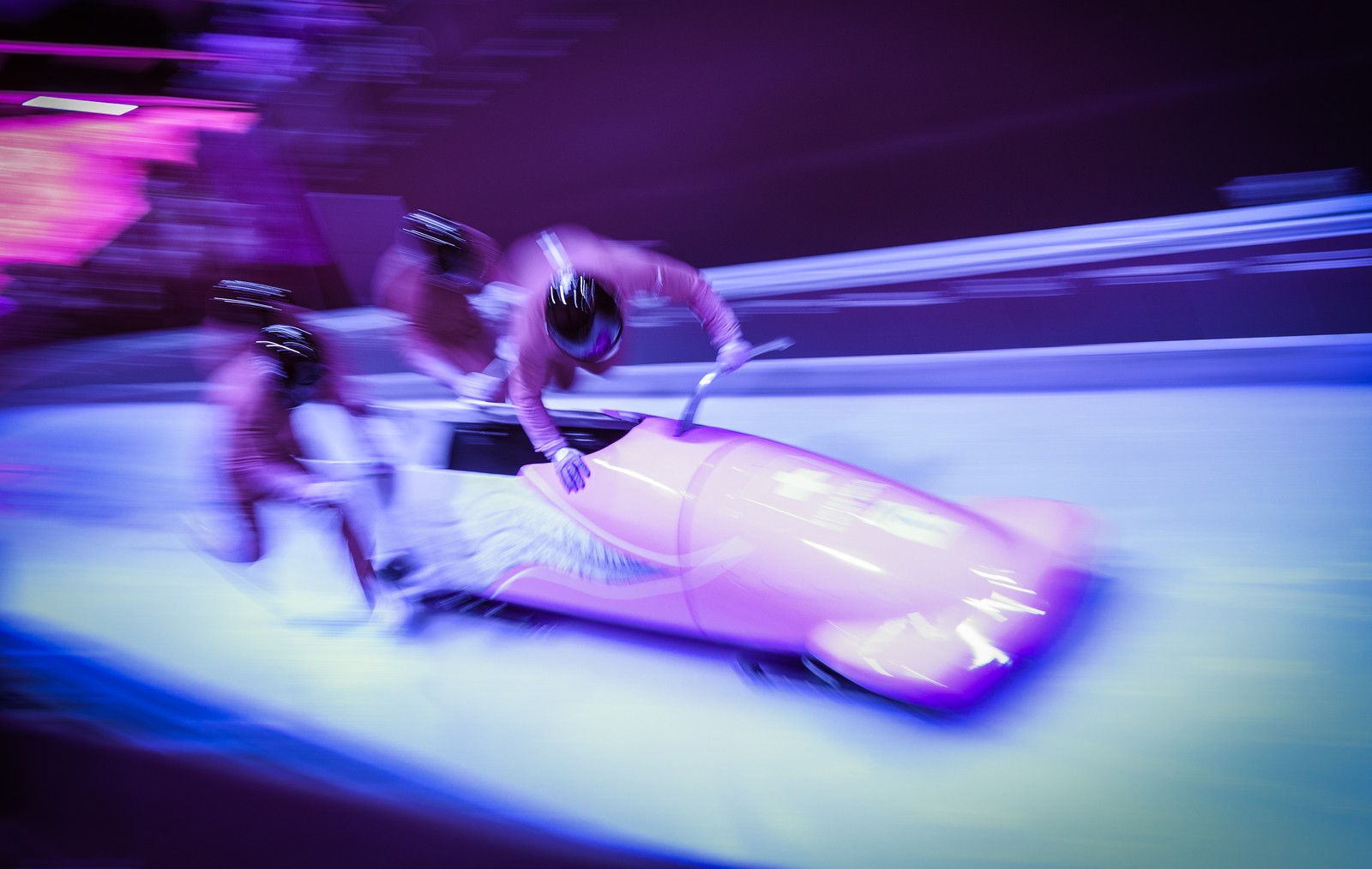 A motion-blurred pink-hued image of three bobsleigh racers running while pushing their sled.