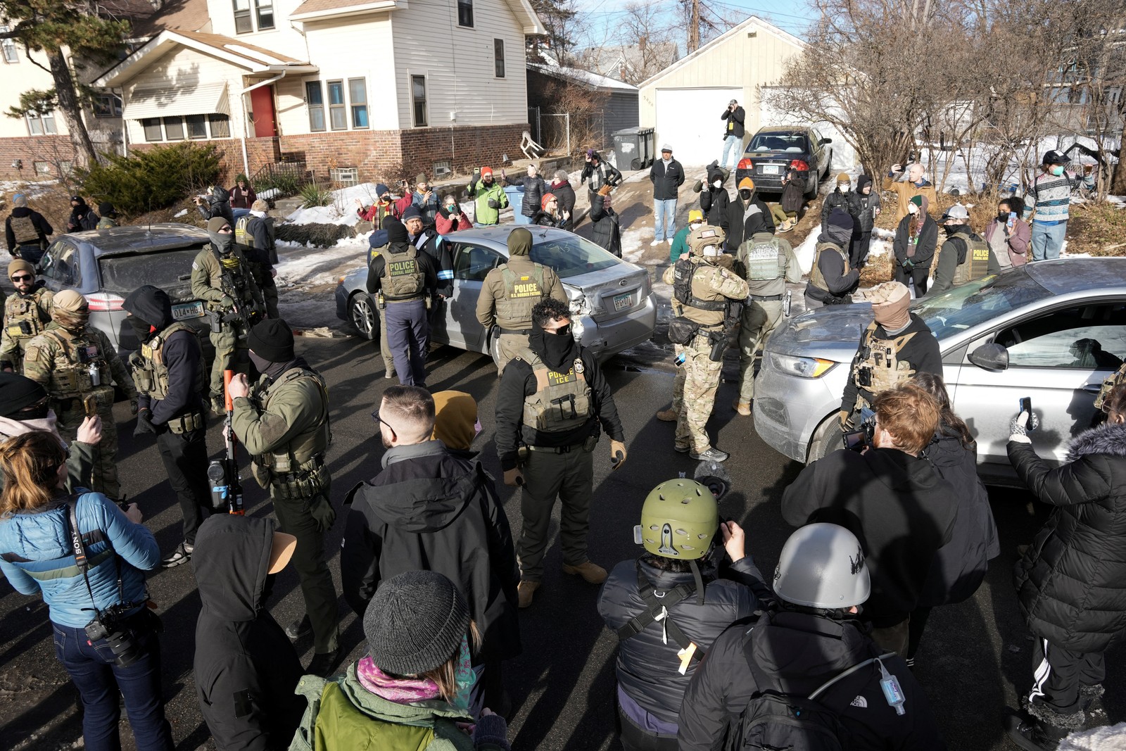 A crowd of onlookers and residents stand on sidewalks, observing more than a dozen masked federal agents standing around a damaged car in a residential neighborhood.