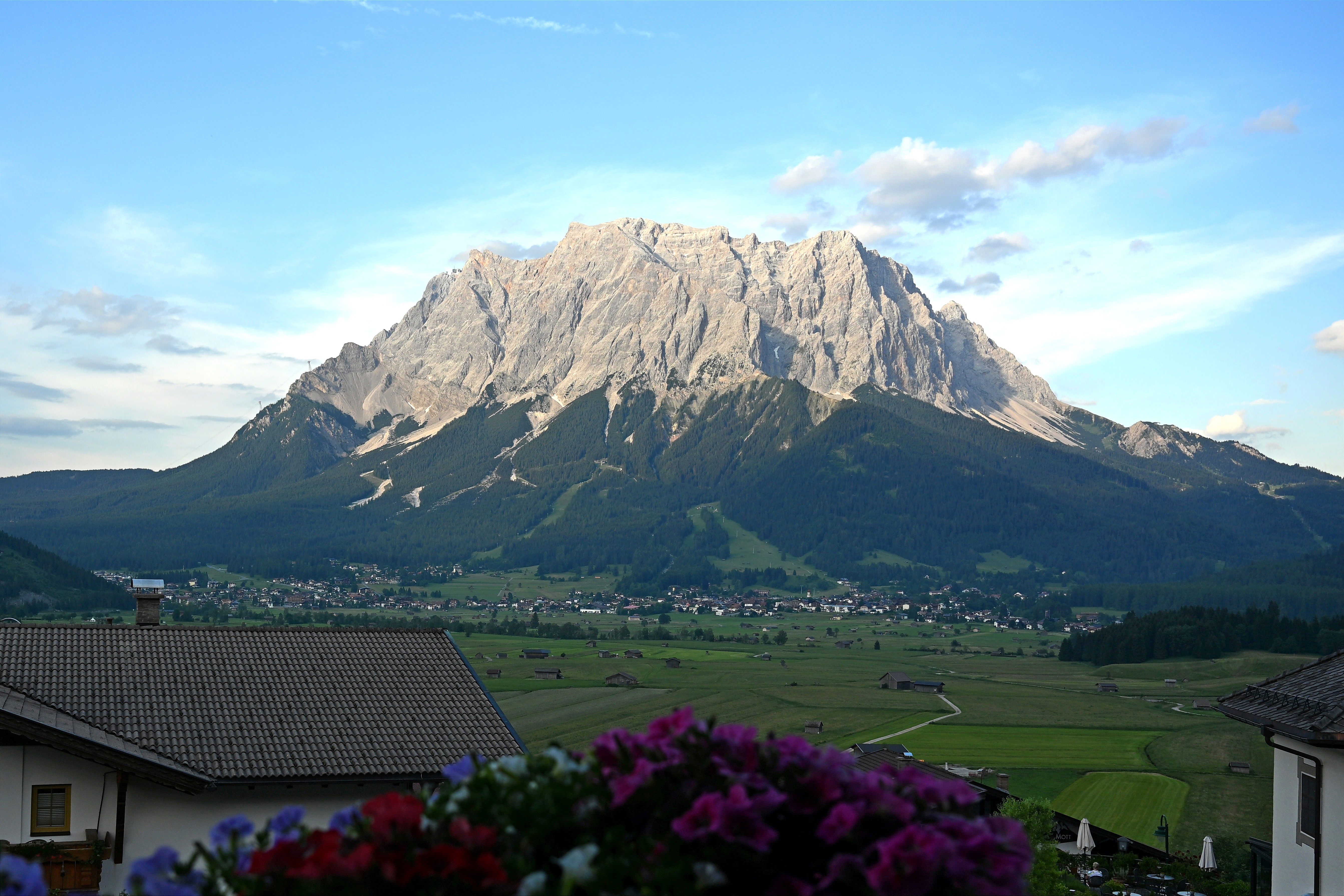 A view of a tall and broad mountain standing above a green valley dotted with houses and farms. 