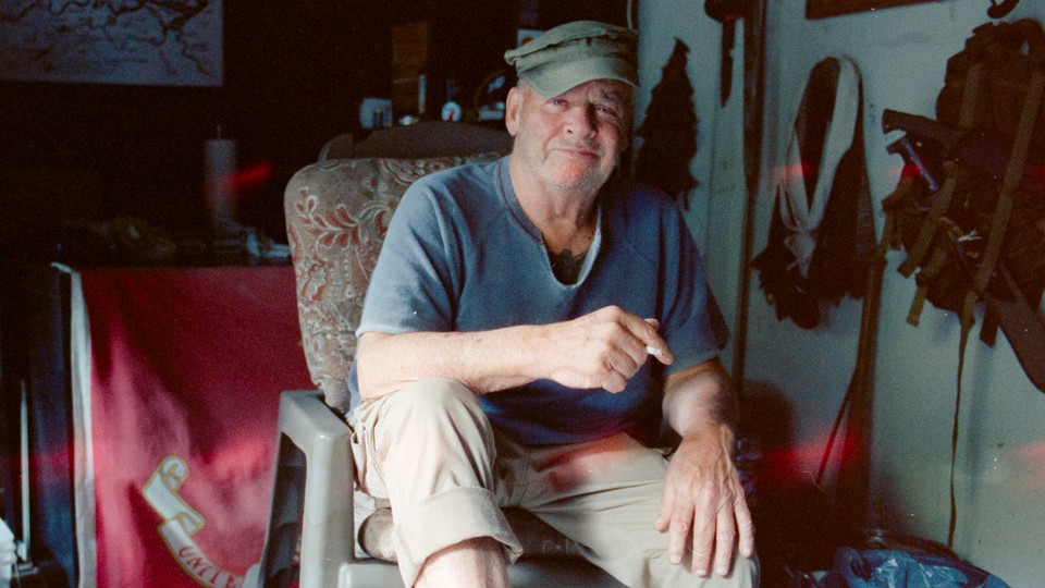 A white man sits in a chair holding a cigarette.