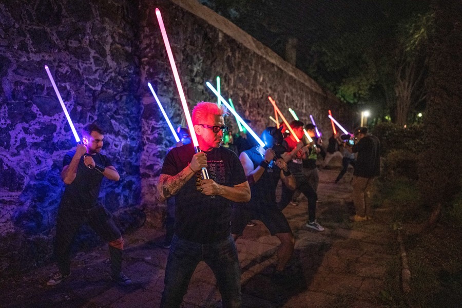 More than a dozen people stand side by side near a wall, holding glowing lightsaber replicas.