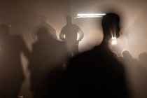 Photograph of black silhouettes of border agents standing with hands on their hips illuminated by a vehicle’s headlights behind them