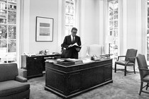 Henry Kissinger standing at a desk in an office with high windows in 1970