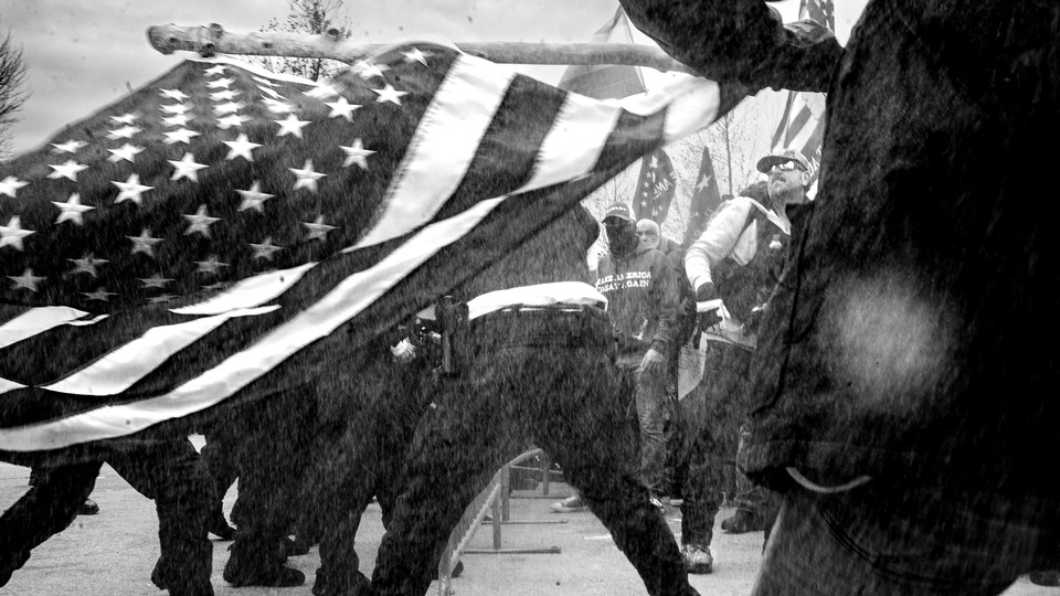 A black-and-white photo of an American flag held sideways in front of a row of cops facing off against men in tactical vests and MAGA sweatshirts.
