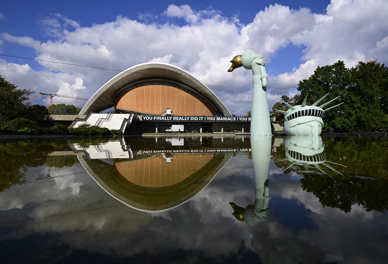 An inflatable replica of the Statue of Liberty sits, mostly-submerged in a pool of water in front of an exhibition hall, with a sign reading 