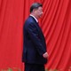 Photographie de Xi Jinping marchant vers la droite, avec de grands rideaux rouges suspendus derrière lui et les feuilles supérieures de petites plantes au premier plan.