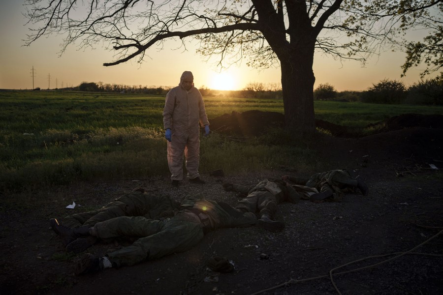 A person clad in protective gear stands beside the bodies of at least four soldiers lying on the ground.