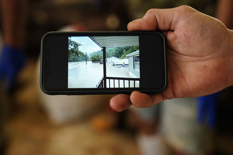 A person holds up their phone showing a photograph of a flooded residential area.