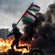 A man holding a Palestinian flag runs in front of a blazing fire.
