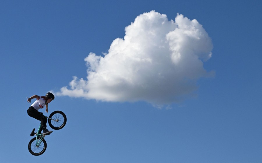 A competitor makes a jump with her BMX bike. A single large cloud floats in the blue sky beyond.