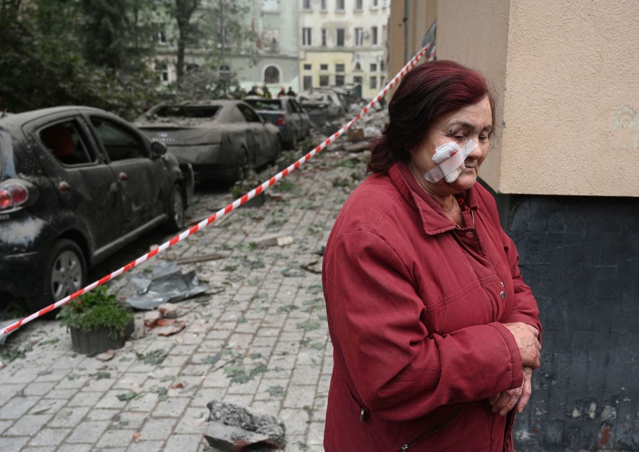 A person stands near debris from a bomb-damaged building, with a bandage on her face.