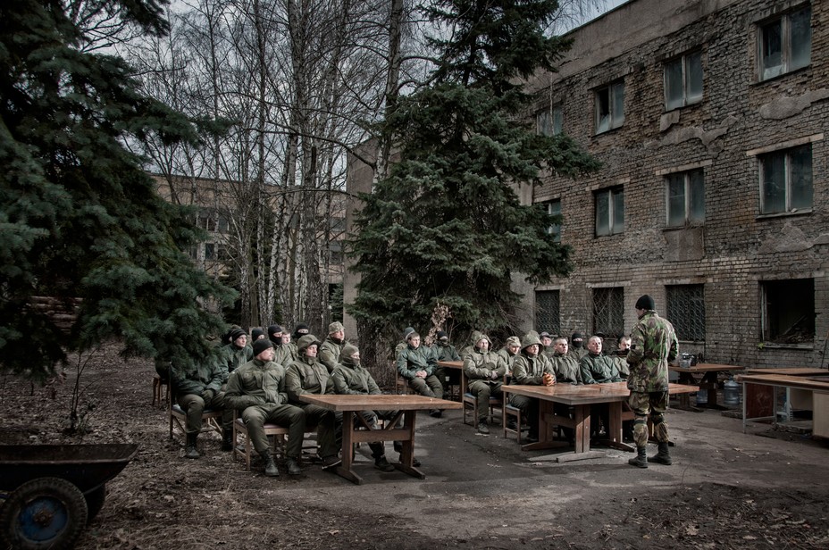 a group of people dressed in fatigues sitting in rows listening to a man talk outside