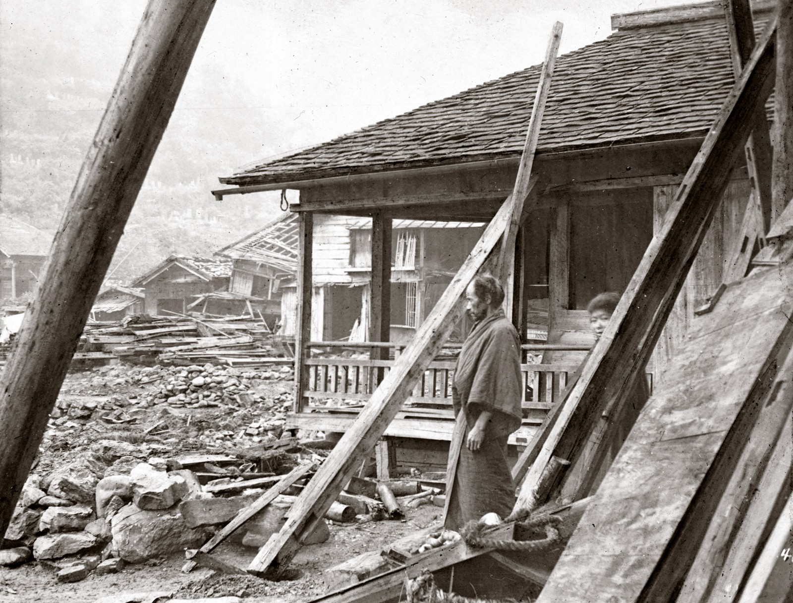 Two people stand outside a damaged house, looking at debris from other earthquake-damaged houses.
