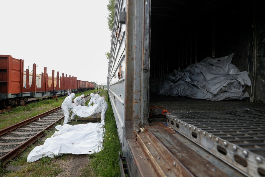 Four people in protective gear lift white body bags from the ground to place them in a nearby railroad car.