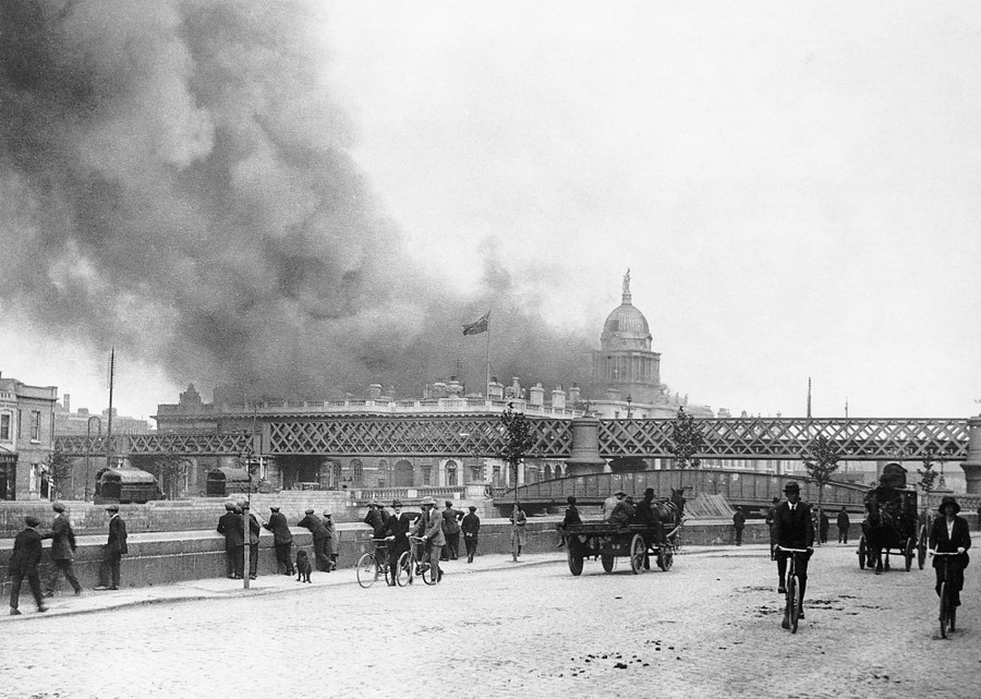 Smoke rises from a large building, as pedestrians watch from a nearby street.