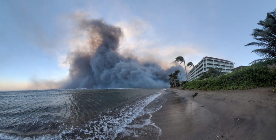 Smoke billows over the ocean, blown from burning structures near a beach.