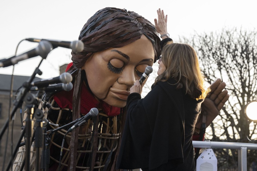A giant puppet of a girl leans down toward a speaker.