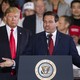 Ron DeSantis stands at a podium, his arms spread open. Donald Trump stands just behind him, on the left.